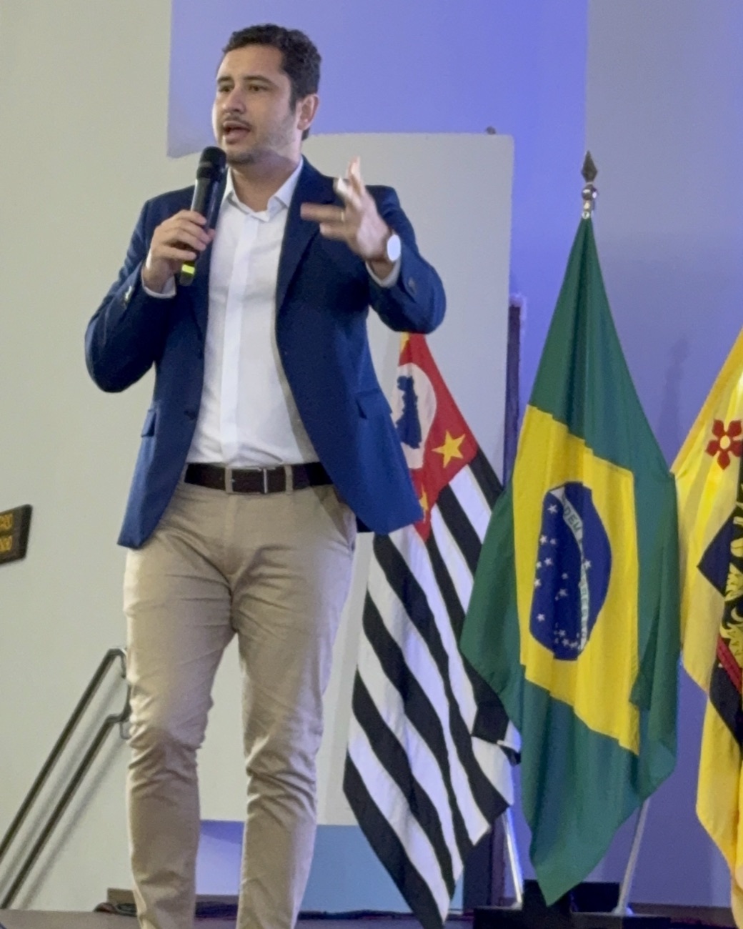 Diego Meloni falando ao microfone numa palestra, com as bandeiras do Brasil e de São Paulo ao fundo